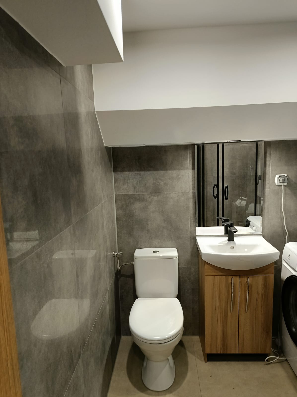 Szara łazienka z toaletą, umywalką na szafce z drewnianym frontem i pralką. Widoczne szare płytki na ścianach i podłodze. Nad umywalką lustro.