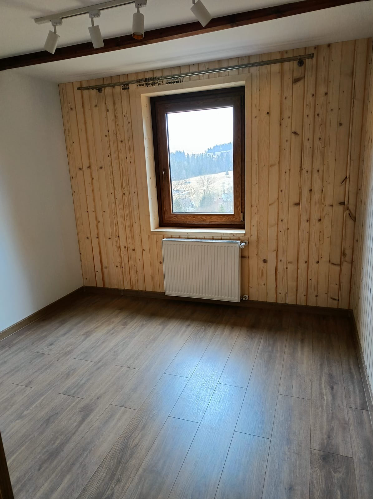 Pokój z drewnianą boazerią, brązowymi panelami podłogowymi i oknem z widokiem na zalesione wzgórza. Grzejnik pod oknem. Oświetlenie sufitowe.