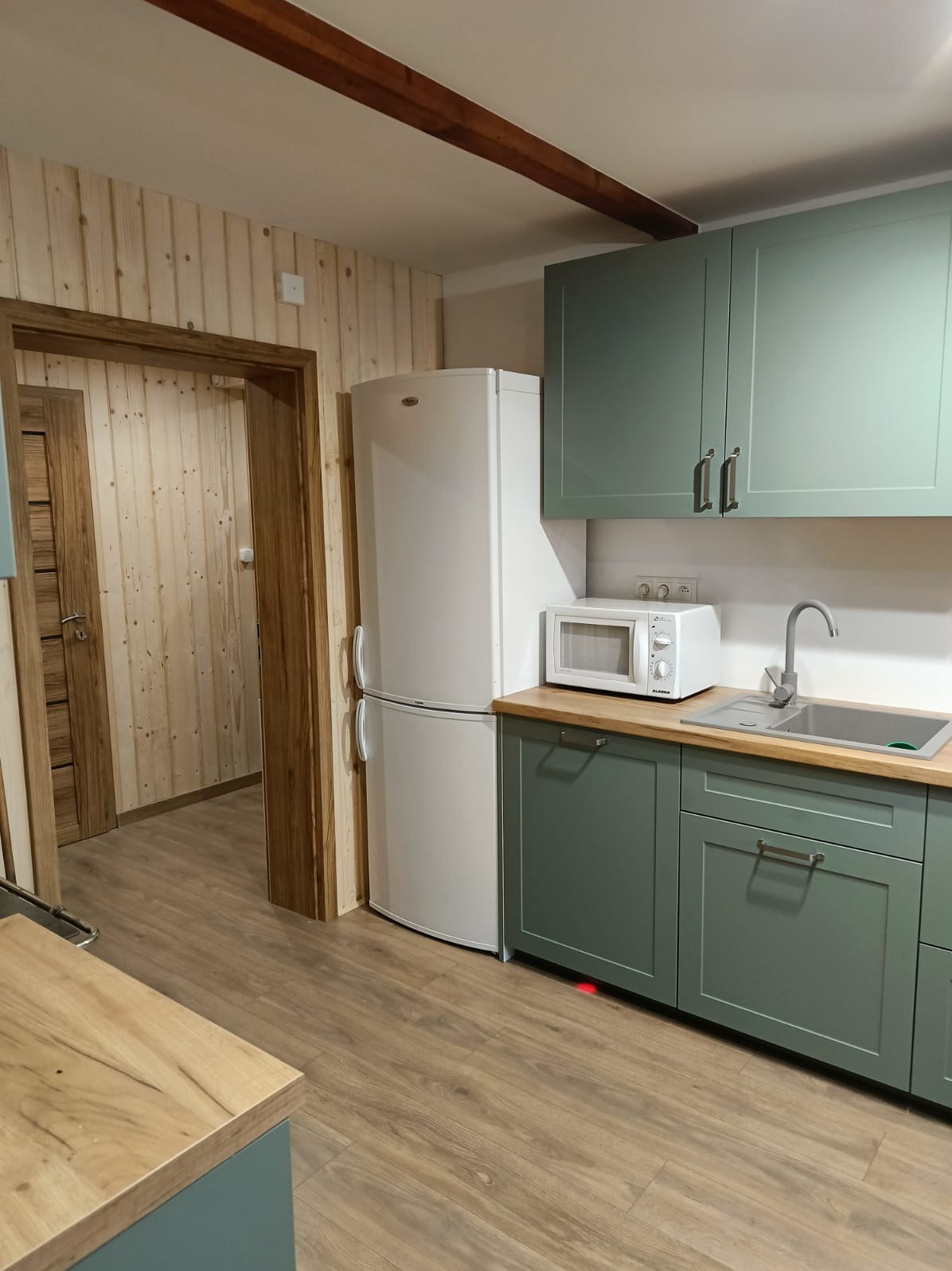 Jasna kuchnia z drewnianą boazerią i zielonymi szafkami. Widoczna lodówka, mikrofalówka i zlew. Drewniane belki na suficie dodają rustykalnego charakteru.