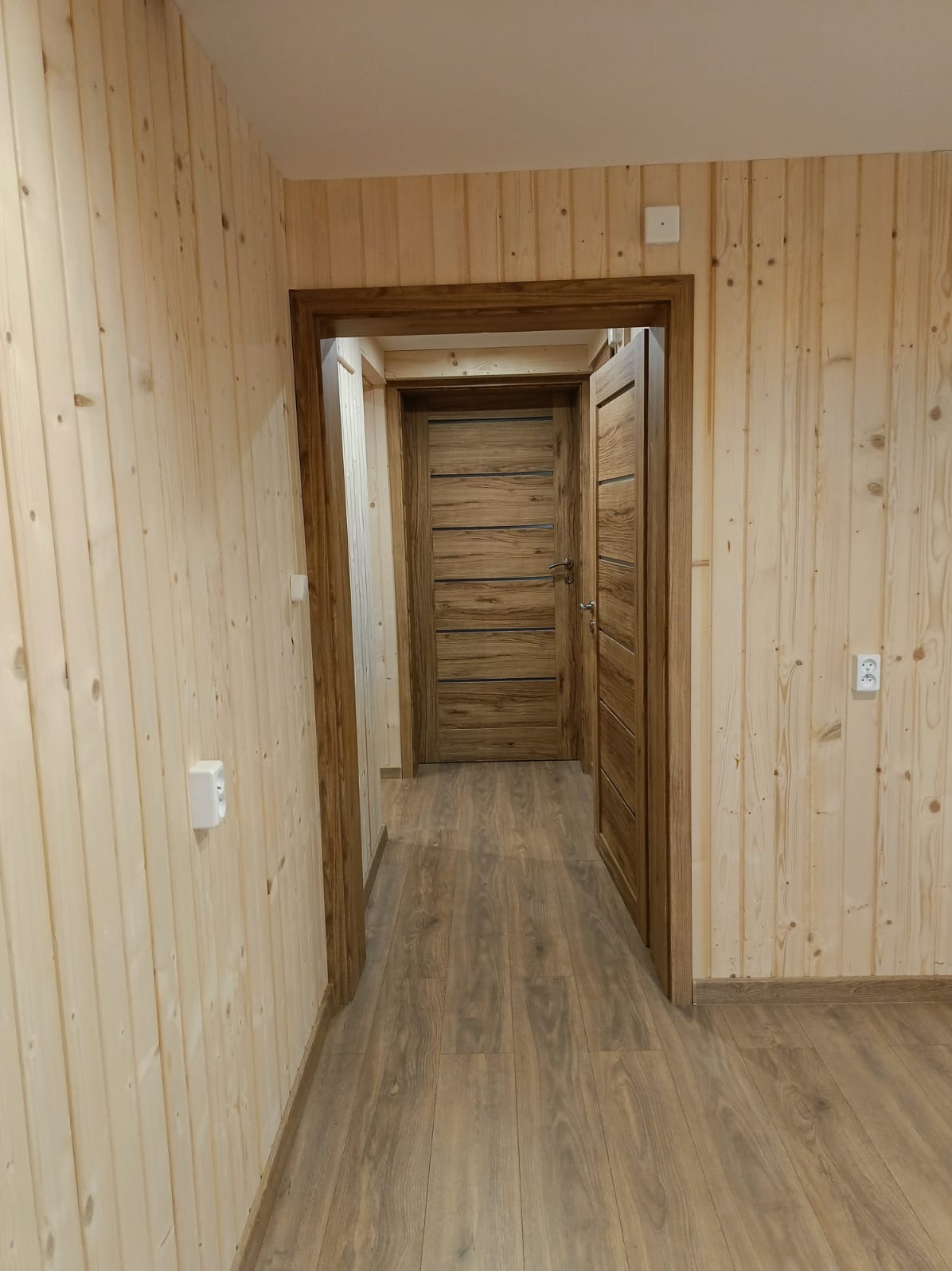 Wnętrze wykończone boazerią i drewnianą podłogą. Widoczne dwoje drewnianych drzwi z ciemniejszymi ościeżnicami. Jasne, naturalne oświetlenie.