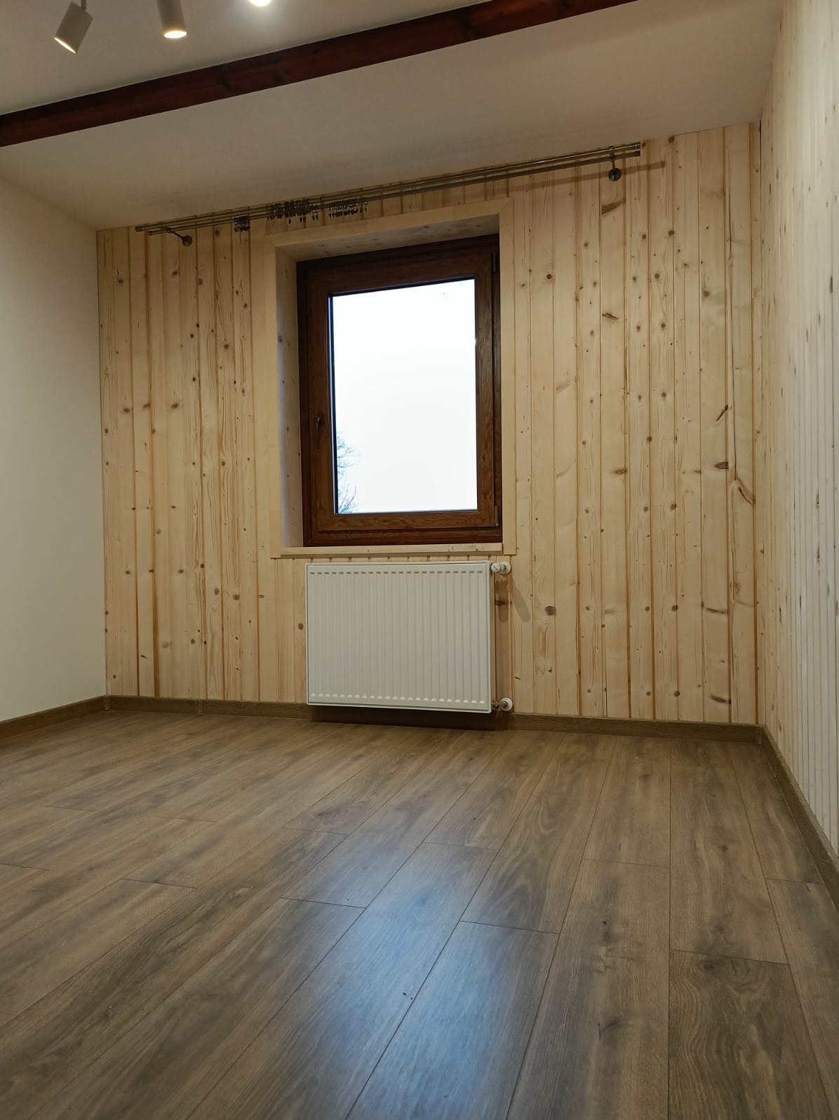 Wnętrze pokoju z drewnianą boazerią na ścianach, brązowym oknem i panelami podłogowymi. Widoczny biały grzejnik pod oknem i karnisz. Ujęcie z niskiego kąta.