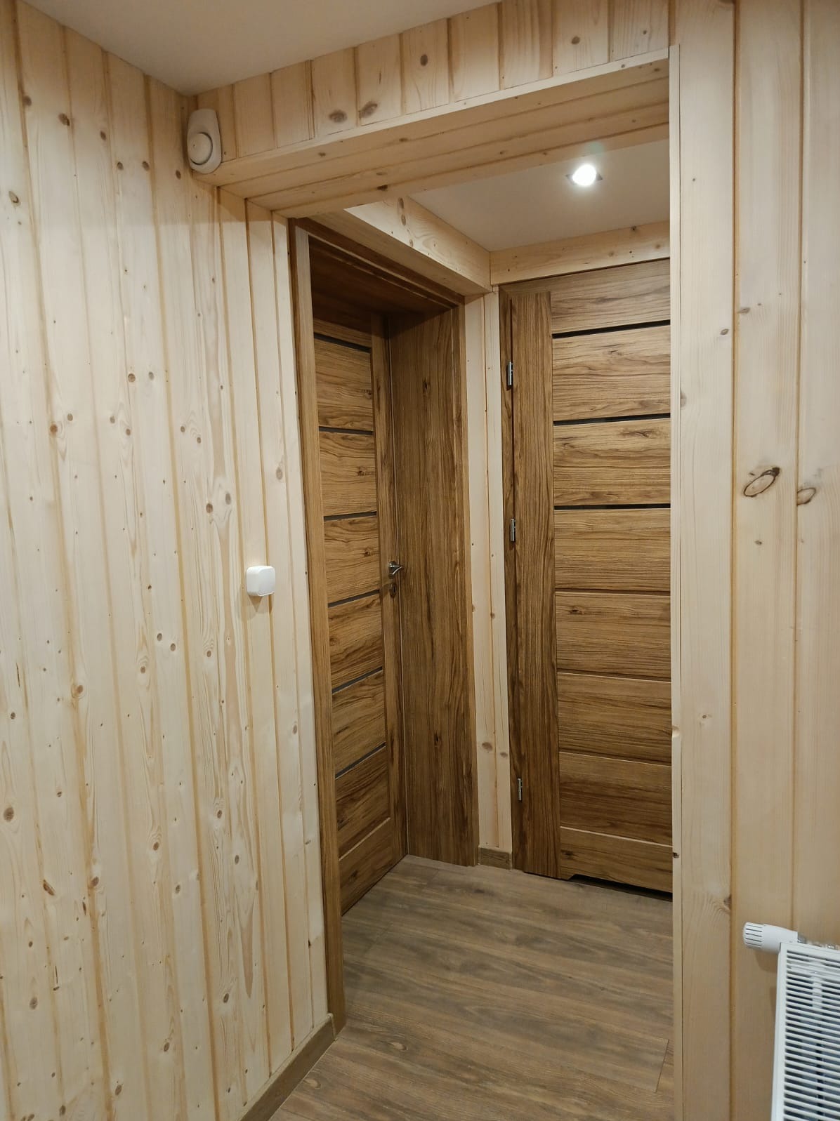 Wnętrze wykończone boazerią i drewnianymi drzwiami z poziomymi panelami. Widoczny fragment grzejnika i detektor dymu. Ciepłe oświetlenie.