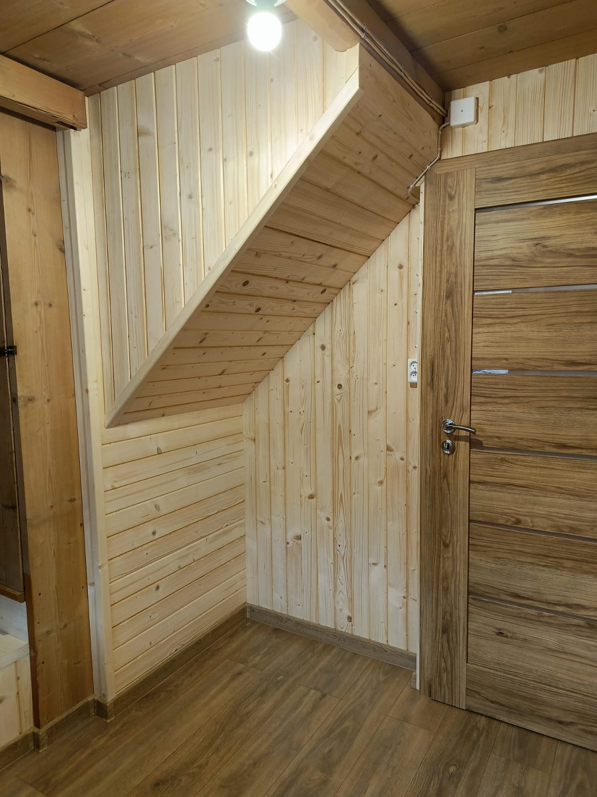 Drewniane wykończenie wnętrza: boazeria ścienna, podbitka schodów i drzwi z drewnianą okleiną. Jasne drewno, ciepłe oświetlenie, widoczne gniazdko elektryczne.