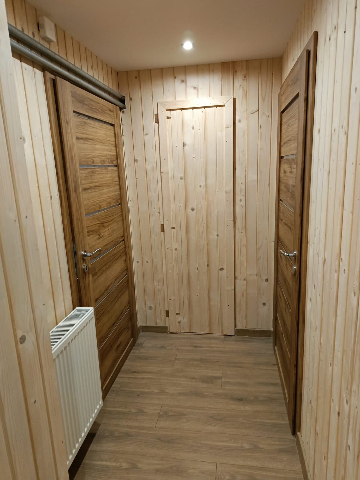 Wnętrze wykończone boazerią z trzema drewnianymi drzwiami, z czego jedne przesuwne. Widoczny grzejnik i podłoga imitująca drewno. Ciepłe oświetlenie.