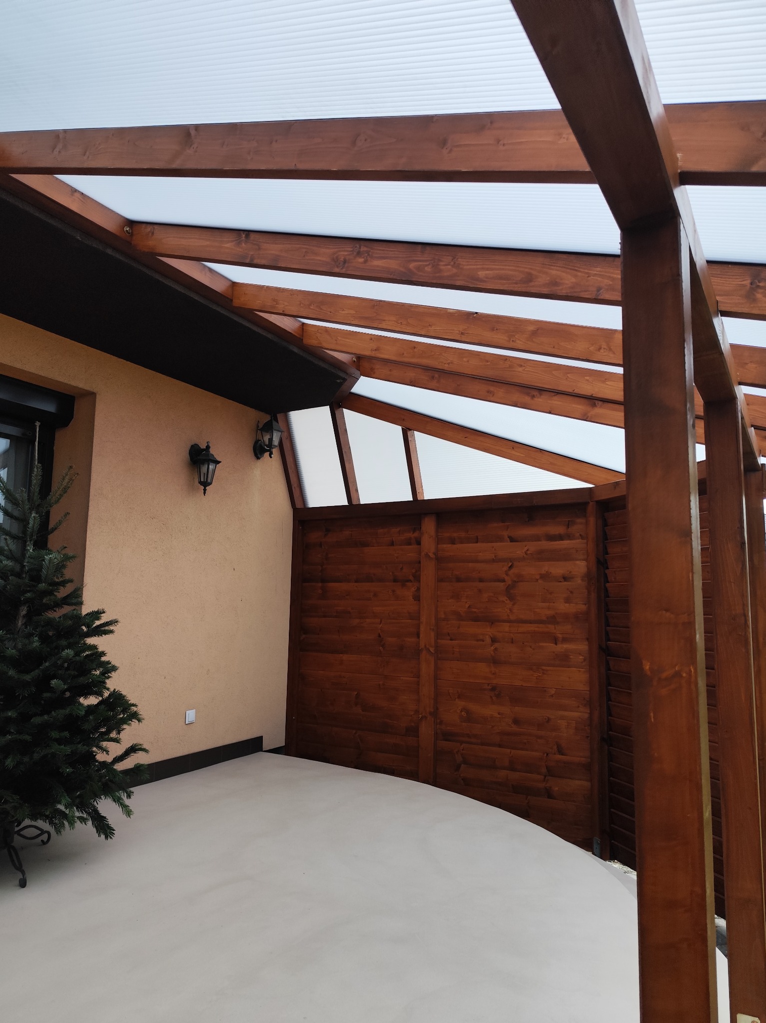 Zadaszona konstrukcja z drewna i przezroczystego tworzywa, przylegająca do budynku. Widoczna drewniana ściana i dekoracyjna choinka. Oświetlenie zewnętrzne.