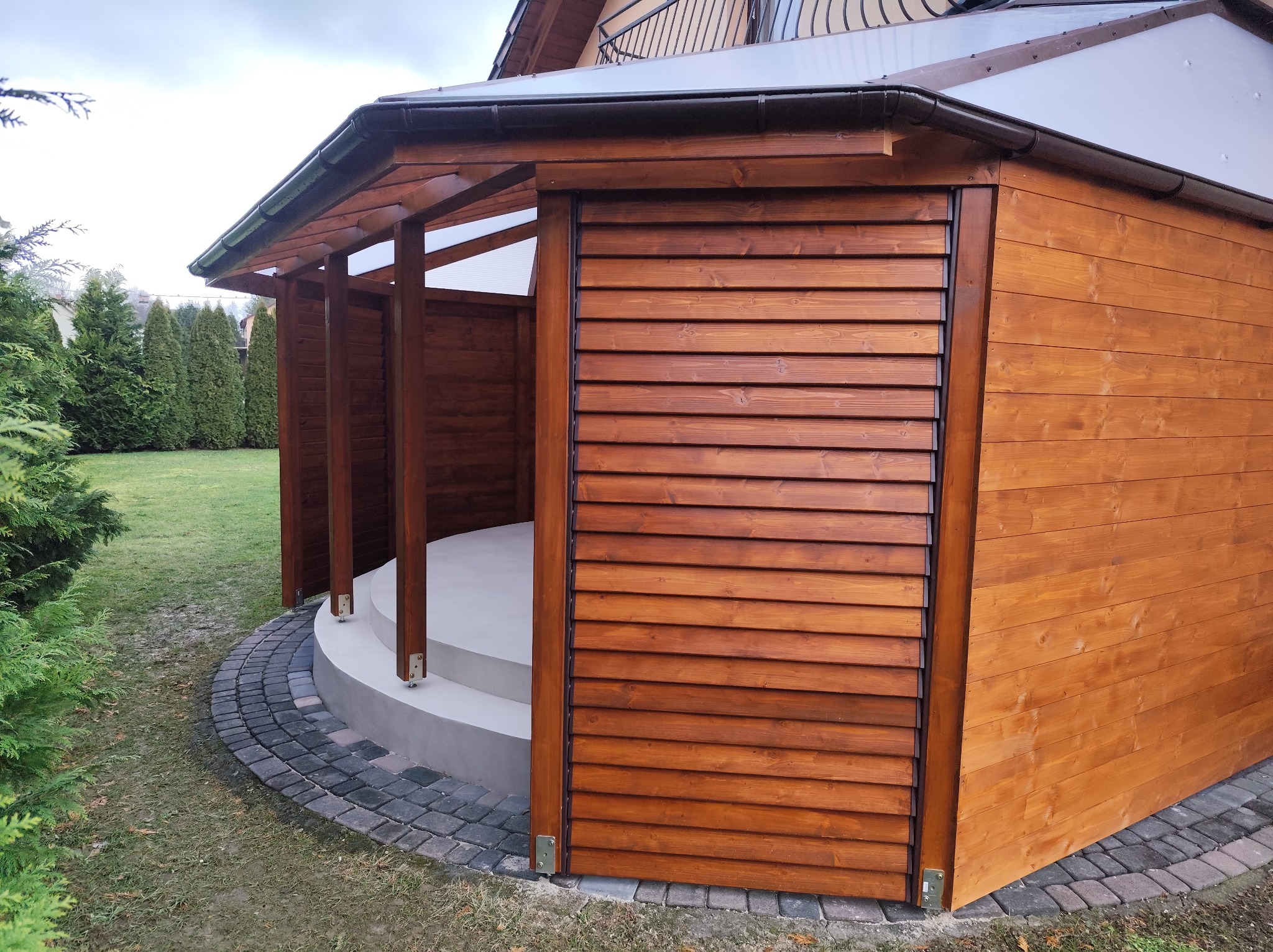 Drewniana altana z lamelami i przezroczystym dachem, otoczona kostką brukową i zielenią. Widoczne detale konstrukcyjne i wykończenie drewna. Ujęcie z boku, w pochmurny dzień.