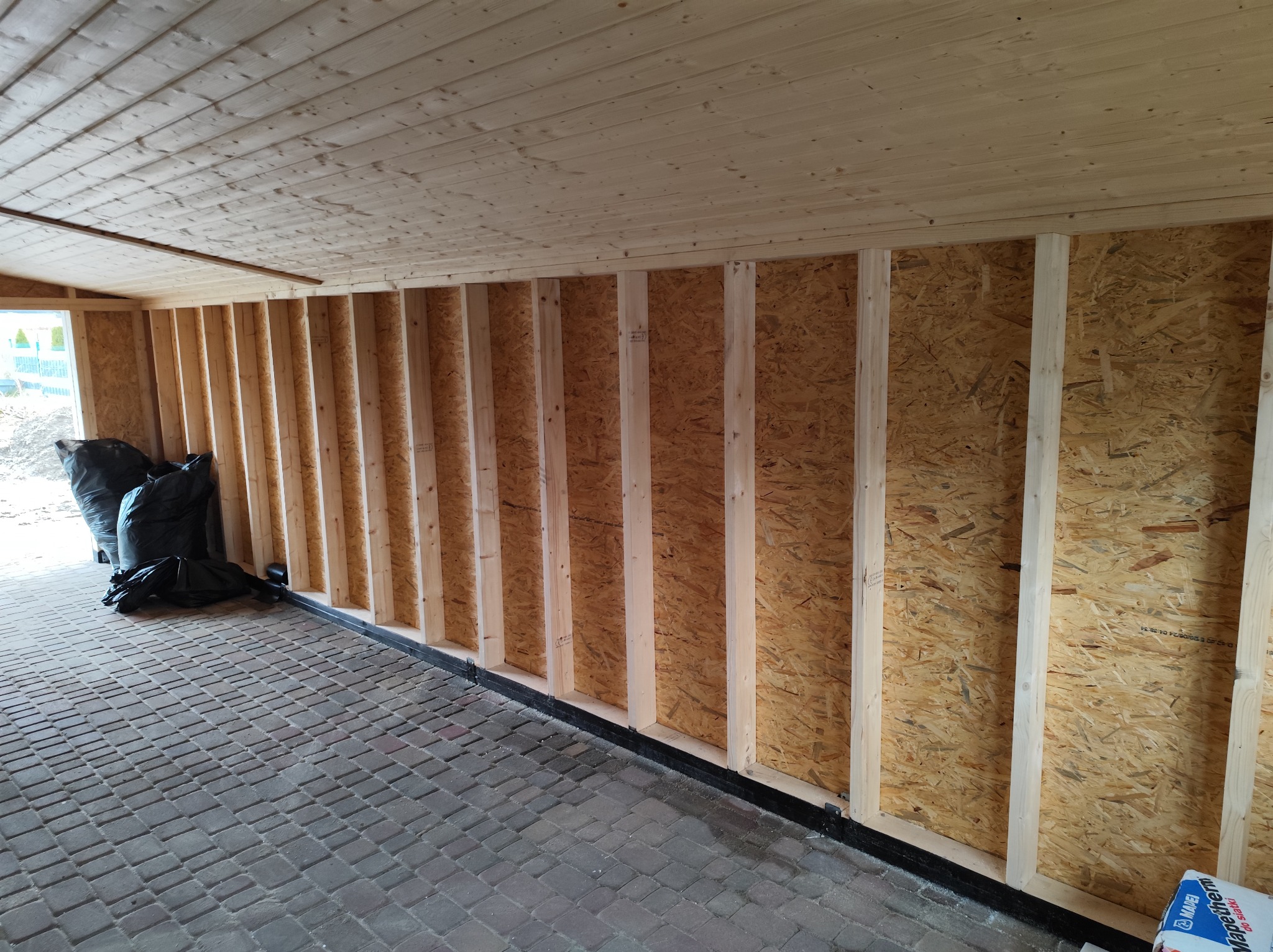 Wnętrze w trakcie budowy domu szkieletowego: drewniana konstrukcja ścian z płyt OSB, drewniany sufit, podłoga z kostki brukowej, czarne worki w rogu.