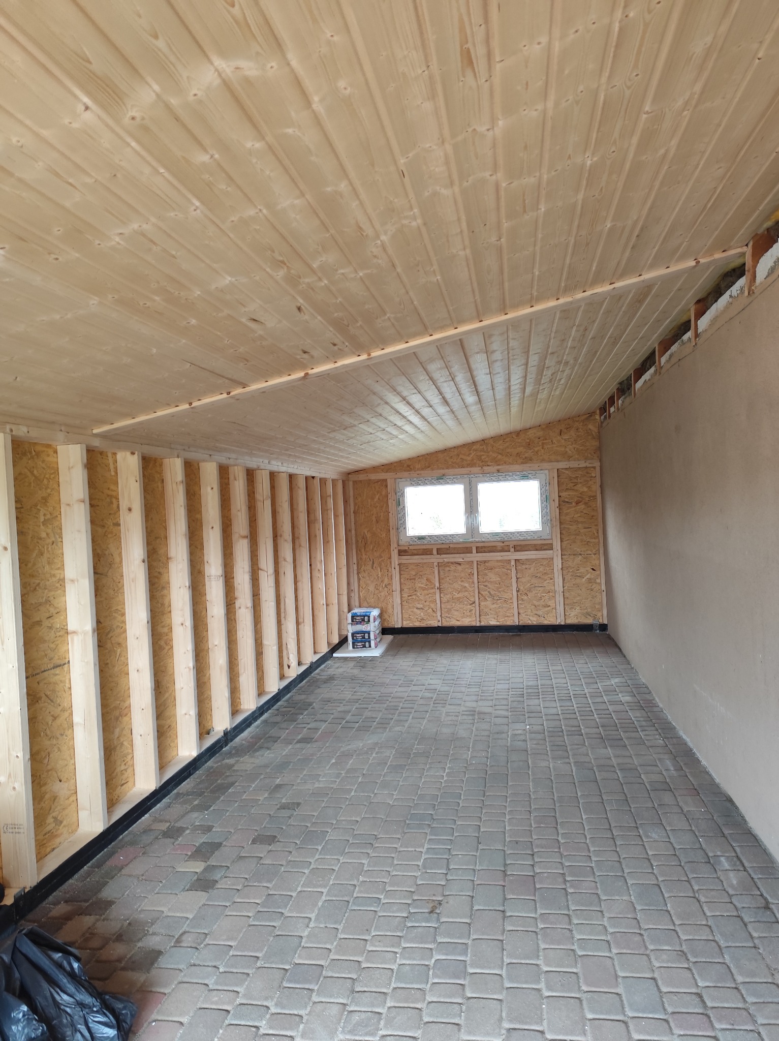 Wnętrze w trakcie budowy: drewniana boazeria na suficie, ściany z płyt OSB z drewnianą konstrukcją, okno i wyłożona kostką podłoga. Widoczne materiały budowlane.