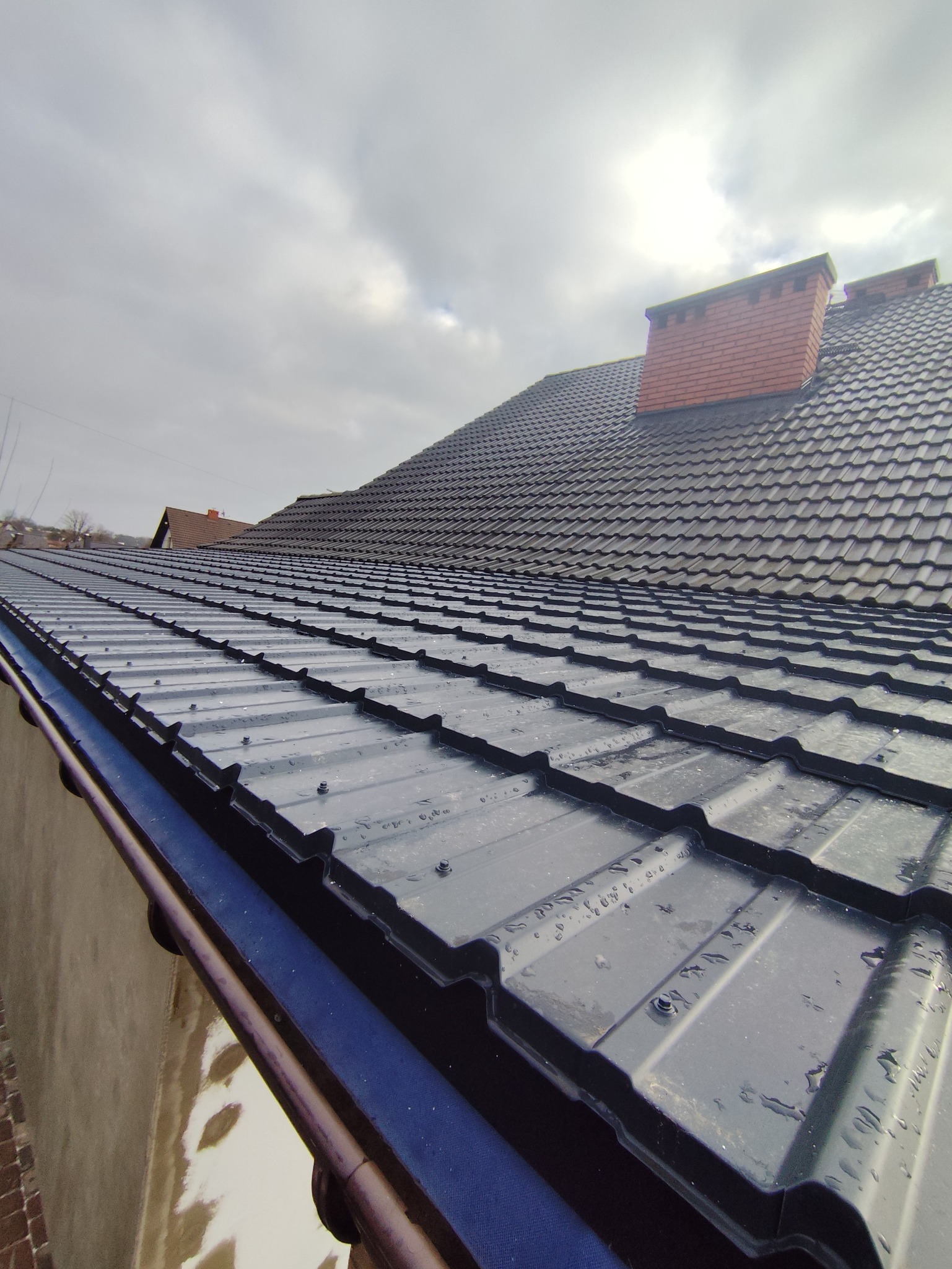 Czarny, metalowy dach z ceglanym kominem w pochmurny dzień. Widoczne krople wody na połaci. Ujęcie z dołu, podkreślające strukturę i detale pokrycia dachowego.