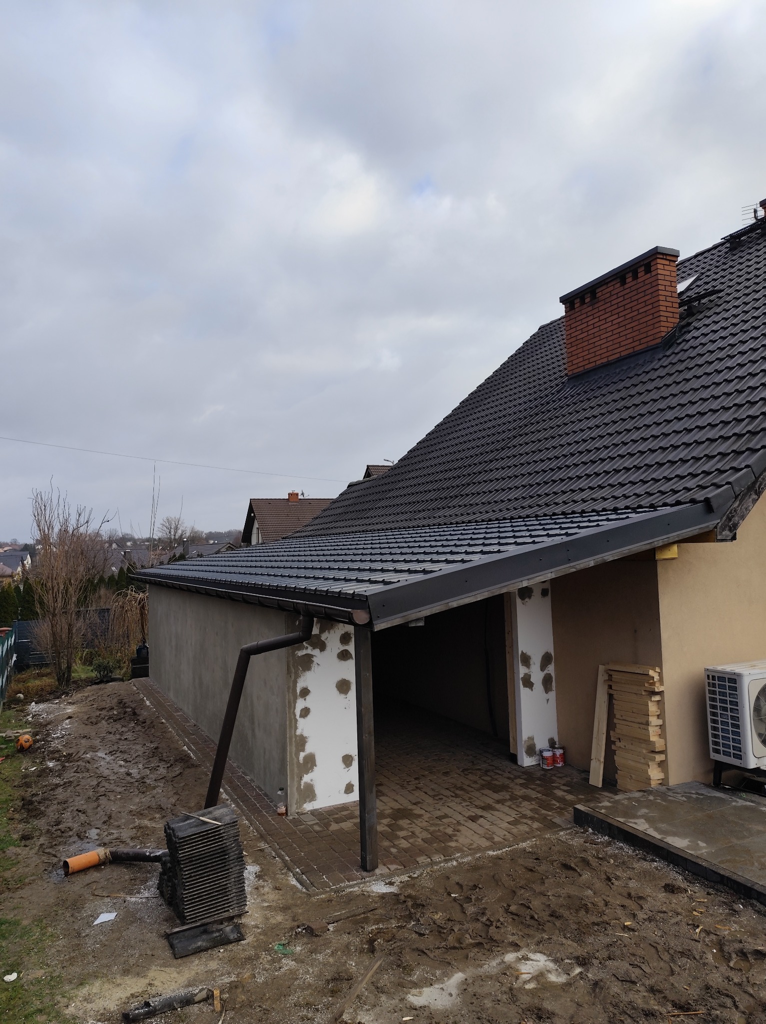 Budowa garażu z dachem z dachówki w Suszcu. Widoczne ściany w trakcie wykończenia, ceglany komin i ułożona kostka brukowa. Prace budowlane w toku.