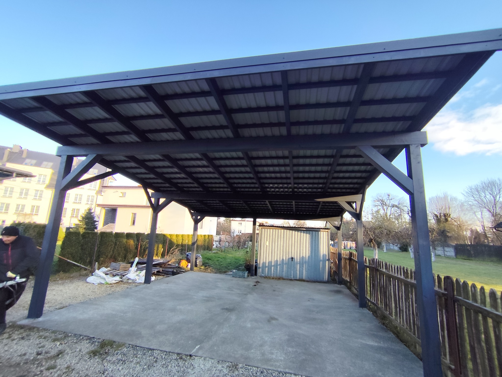 Nowoczesna, ciemna wiata garażowa z metalowym dachem i drewnianą konstrukcją, na betonowej wylewce. W tle budynki mieszkalne i zielony ogród.