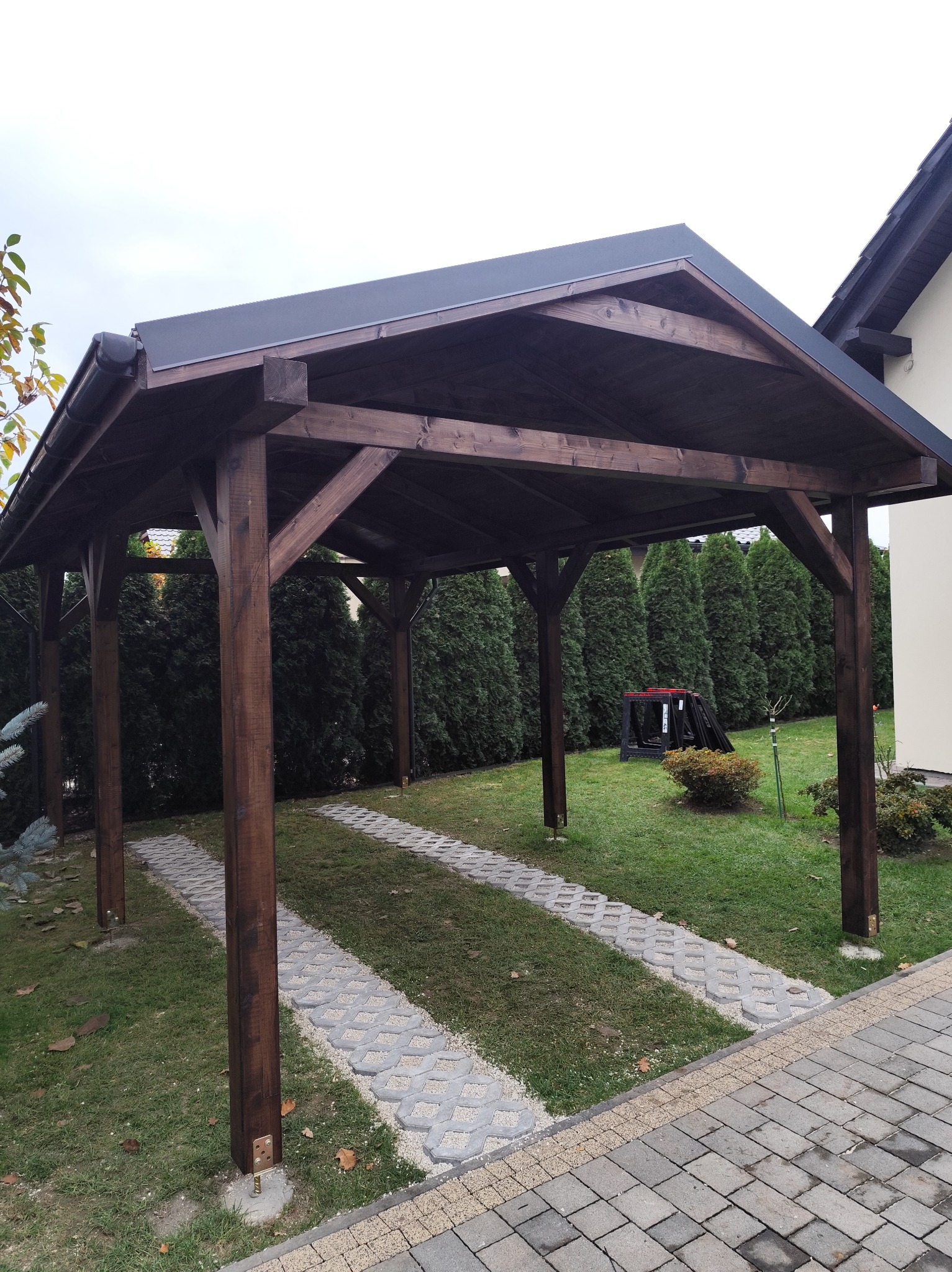 Ciemnobrązowa, drewniana wiata garażowa z płaskim dachem na posesji z trawnikiem i ścieżką z ażurowych płyt. W tle rząd zielonych tuj i fragment domu.