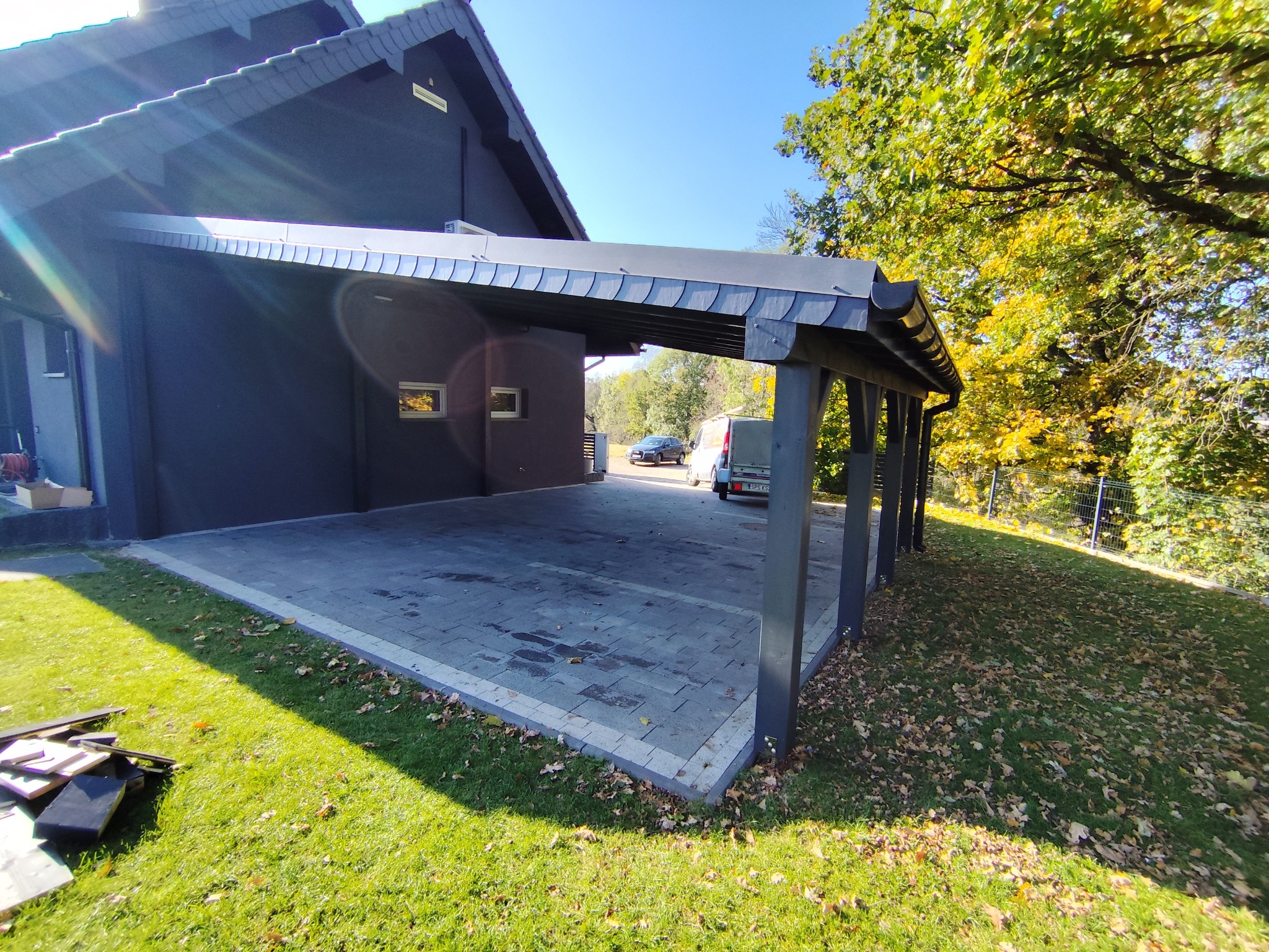 Nowoczesna wiata garażowa z ciemną, płaską dachówką, wsparta na solidnych, szarych słupach. Kostka brukowa w odcieniach szarości. Dom w tle, zieleń drzew.