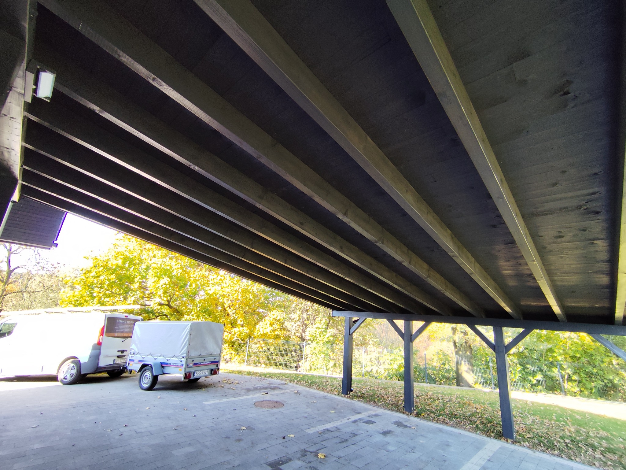 Drewniana pergola z widoczną konstrukcją nośną i zadaszeniem, obok zaparkowany samochód dostawczy i przyczepka. Jesienne otoczenie z drzewami w tle.