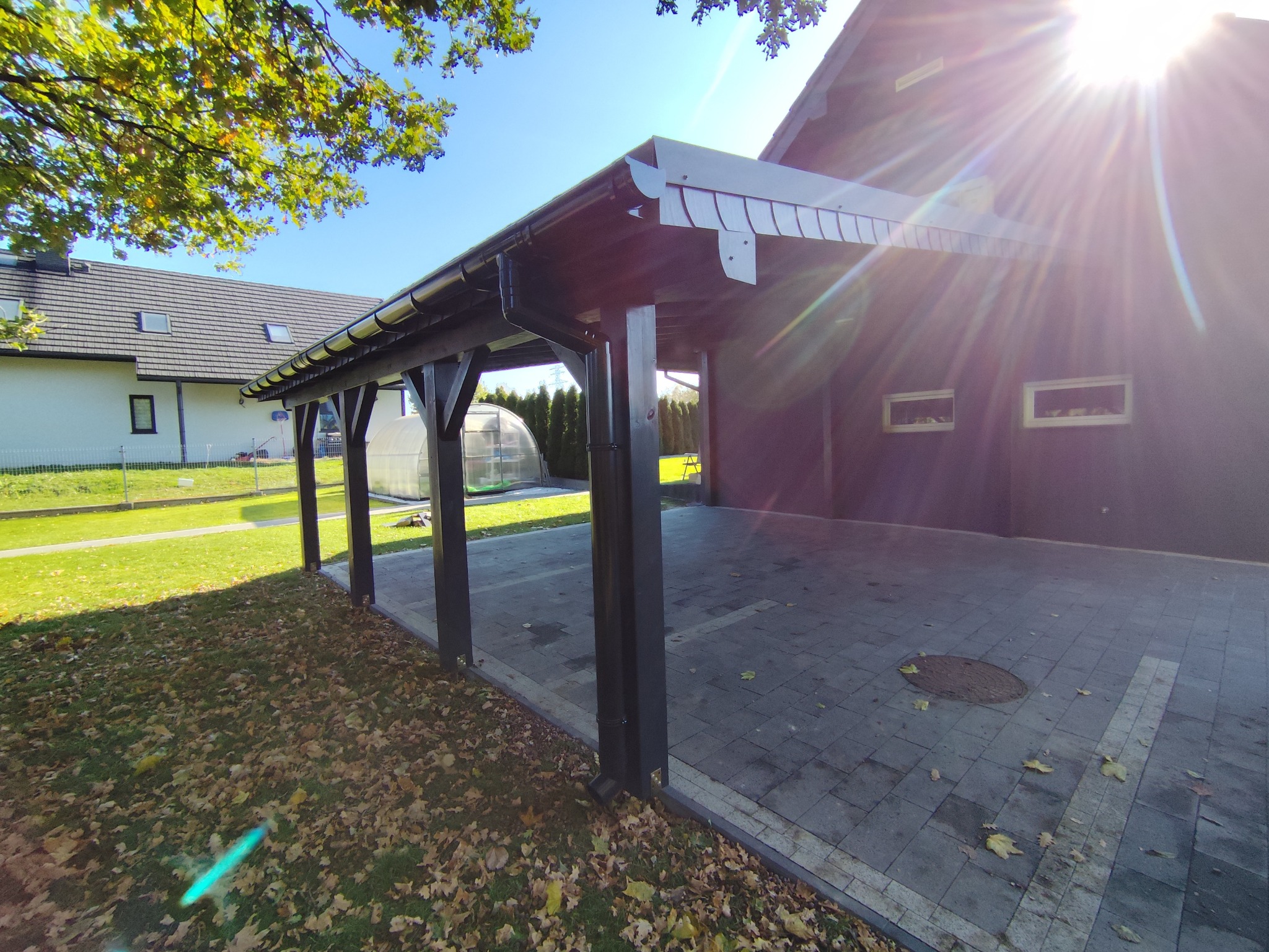 Nowoczesna, grafitowa wiata garażowa z kostki brukowej, z widocznym systemem rynnowym i elementami drewnianymi, w słoneczny dzień. W tle dom z ogrodem.