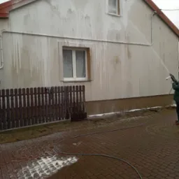 Czyszczenie elewacji domu jednorodzinnego za pomocą myjki ciśnieniowej, widoczne zacieki spływającej wody i piany, częściowo oczyszczona ściana, kostka brukowa z resztkami piany.