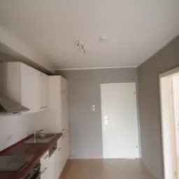 Widok na nową, jasną kuchnię z białymi szafkami, brązowym blatem i płytkami podłogowymi w odcieniach beżu, szare ściany i białe drzwi.