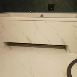 Nowoczesna łazienka z wanną obudowaną płytami imitującymi marmur i zielonymi płytkami ułożonymi w jodełkę na ścianie. Widoczny fragment podłogi wyłożonej płytkami w podobnym stylu do obudowy wanny...