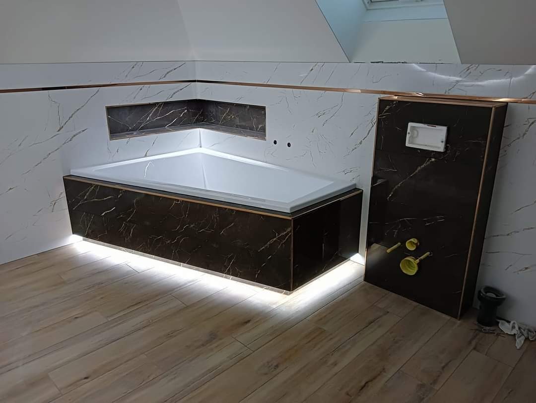 Nowoczesna łazienka z narożną wanną obudowaną panelami imitującymi marmur, podświetleniem LED i płytkami ściennymi z motywem marmuru oraz drewnopodobną podłogą.