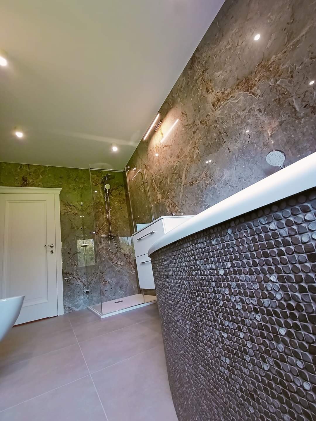 Nowoczesna łazienka z kabiną prysznicową typu walk-in, umywalką w zabudowie i wanną wykończoną mozaiką, ściany wyłożone płytkami imitującymi marmur.