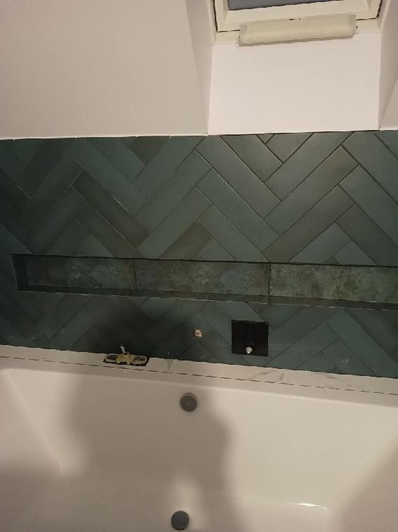 Niebiesko-zielone płytki ułożone w jodełkę nad wanną, z wnęką wykończoną płytkami i czarnym przełącznikiem. Widoczna biała wanna z dwoma odpływami i okno dachowe w tle.