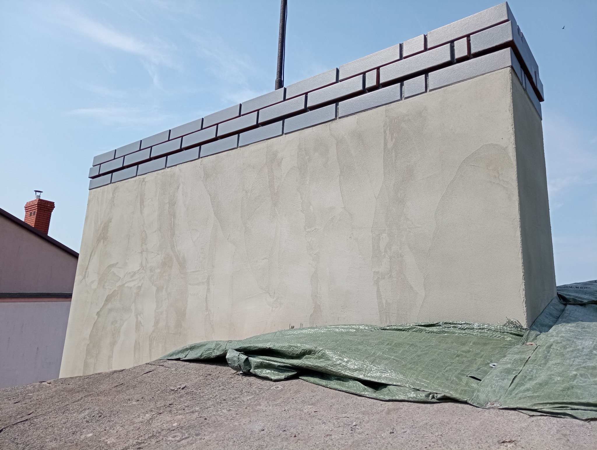 Otynkowany komin z wykończeniem z szarych cegieł, zabezpieczony zieloną plandeką, widok z dołu na tle błękitnego nieba.