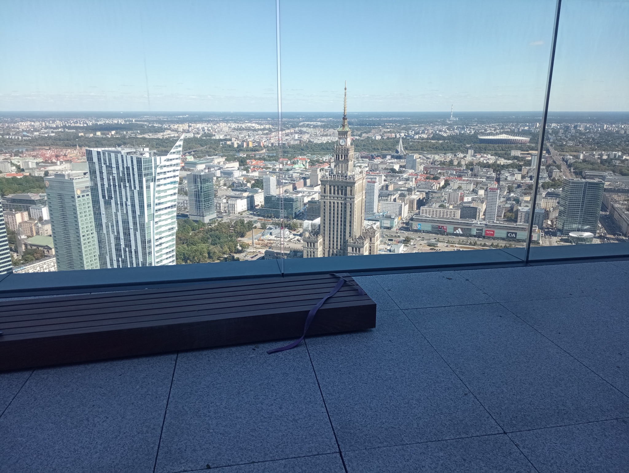 Widok na panoramę Warszawy z wysokiego budynku, na pierwszym planie szare płytki podłogowe i drewniana ławka z fioletową wstążką.