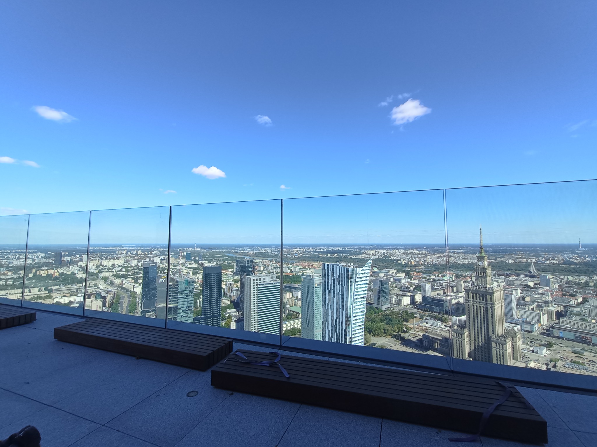 Widok na panoramę miasta z tarasu widokowego z przeszkloną balustradą i drewnianymi ławkami na pierwszym planie, błękitne niebo z pojedynczymi obłokami w tle.