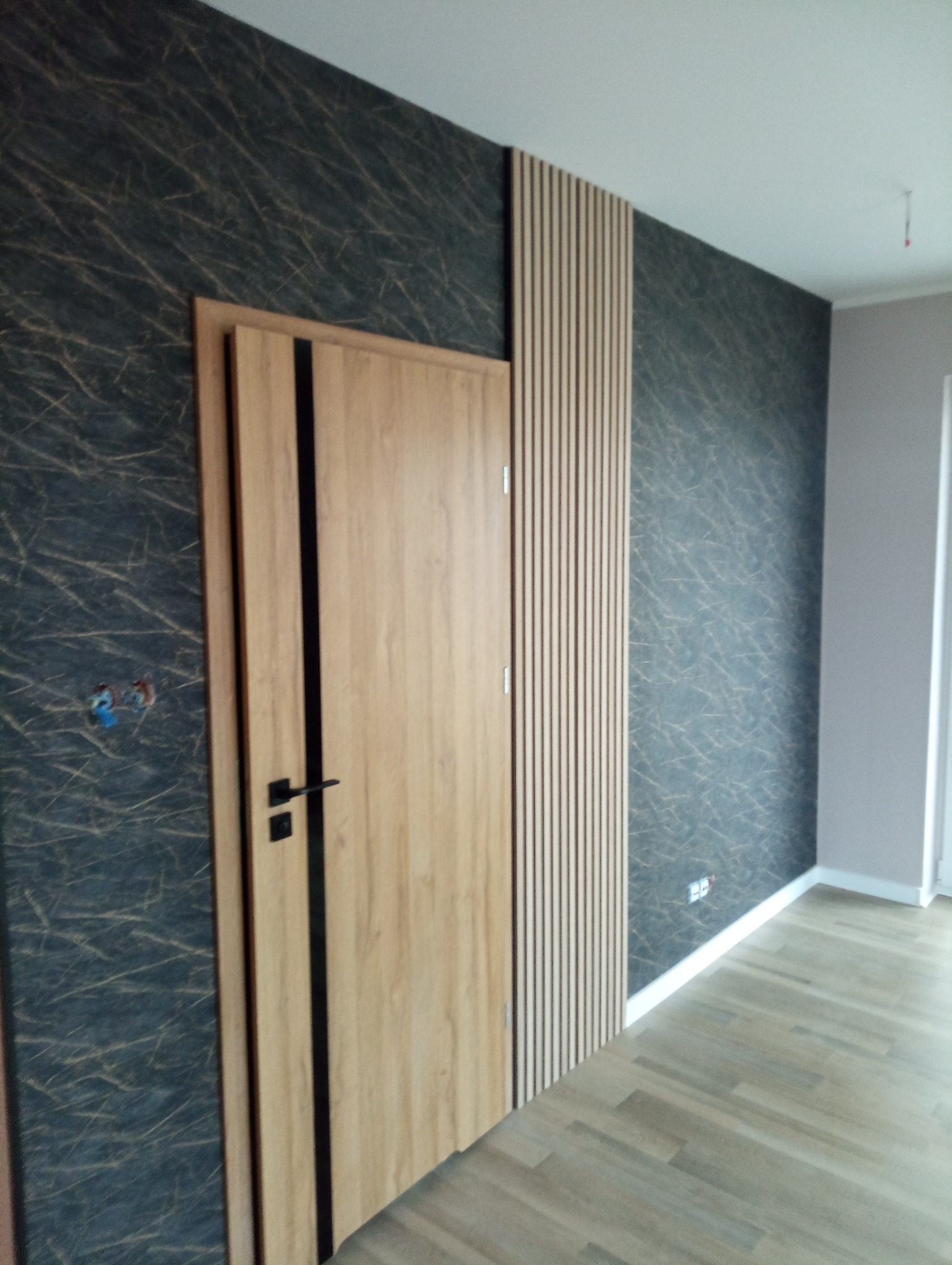 Nowoczesne wykończenie wnętrza: drewniana okleina drzwi z czarnymi akcentami, dekoracyjna ściana z motywem marmuru i pionowe panele ścienne z drewna.