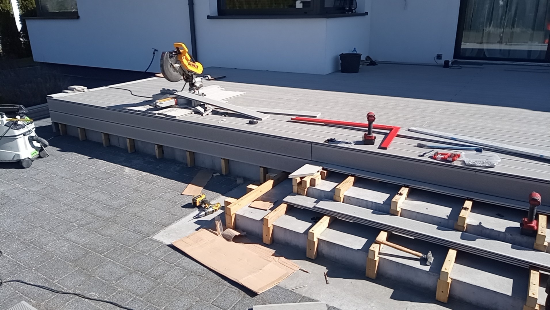 Budowa tarasu kompozytowego na betonowej podstawie z użyciem piły ukośnicy DeWalt, w tle biały budynek z dużym oknem.