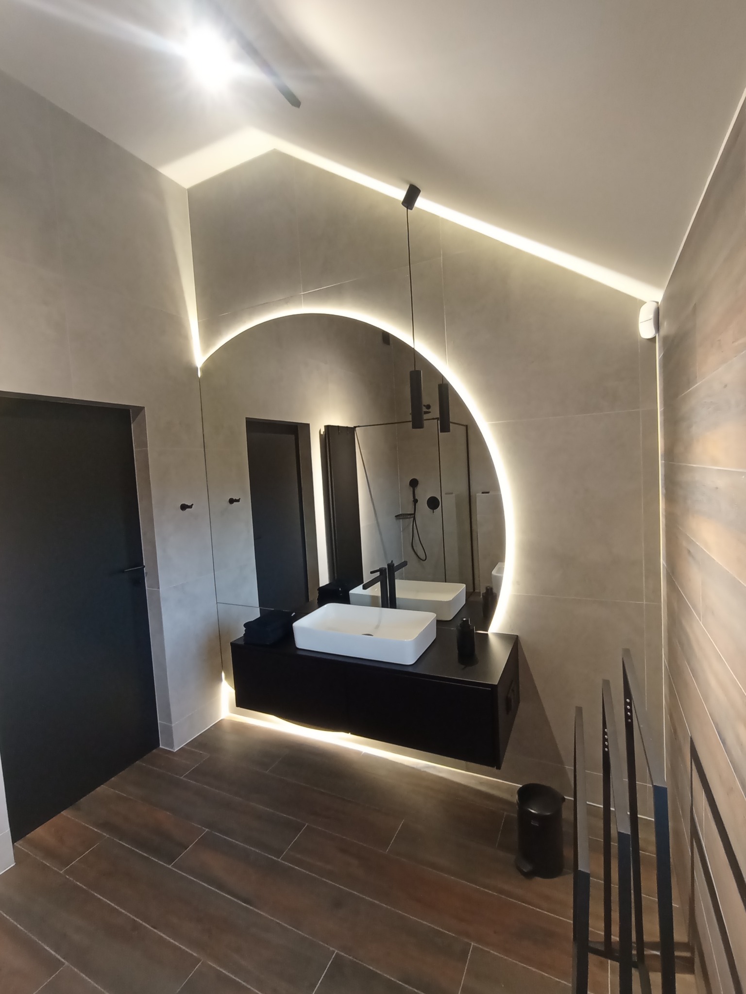 Nowoczesna łazienka z podświetlanym okrągłym lustrem, czarną szafką pod umywalką i brązowymi płytkami podłogowymi imitującymi drewno. Widoczna kabina prysznicowa w odbiciu lustra.