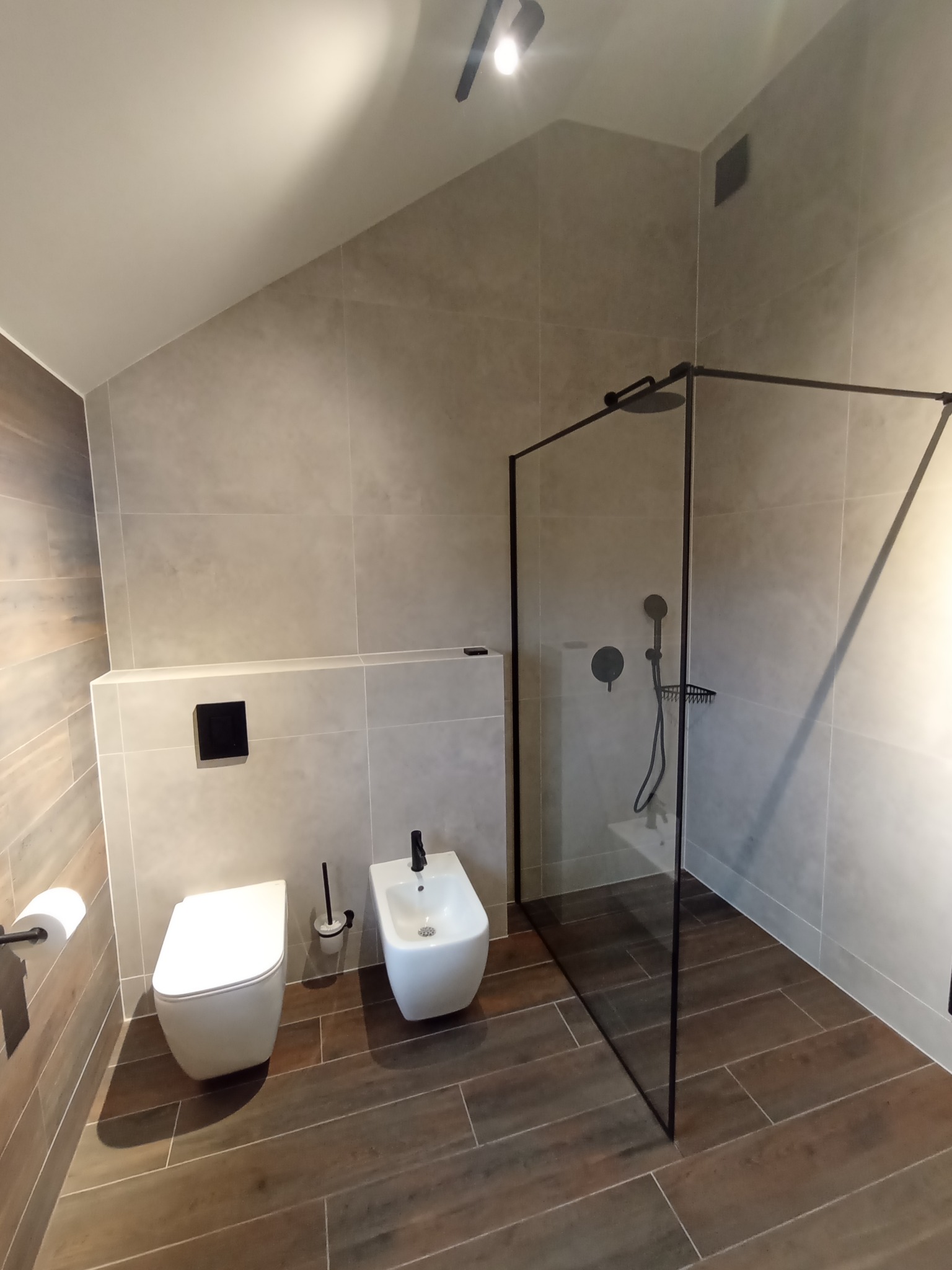 Nowoczesna łazienka z kabiną prysznicową typu walk-in z czarnymi ramami, wiszącą toaletą i bidetem, wykończona płytkami w odcieniach szarości i brązu.
