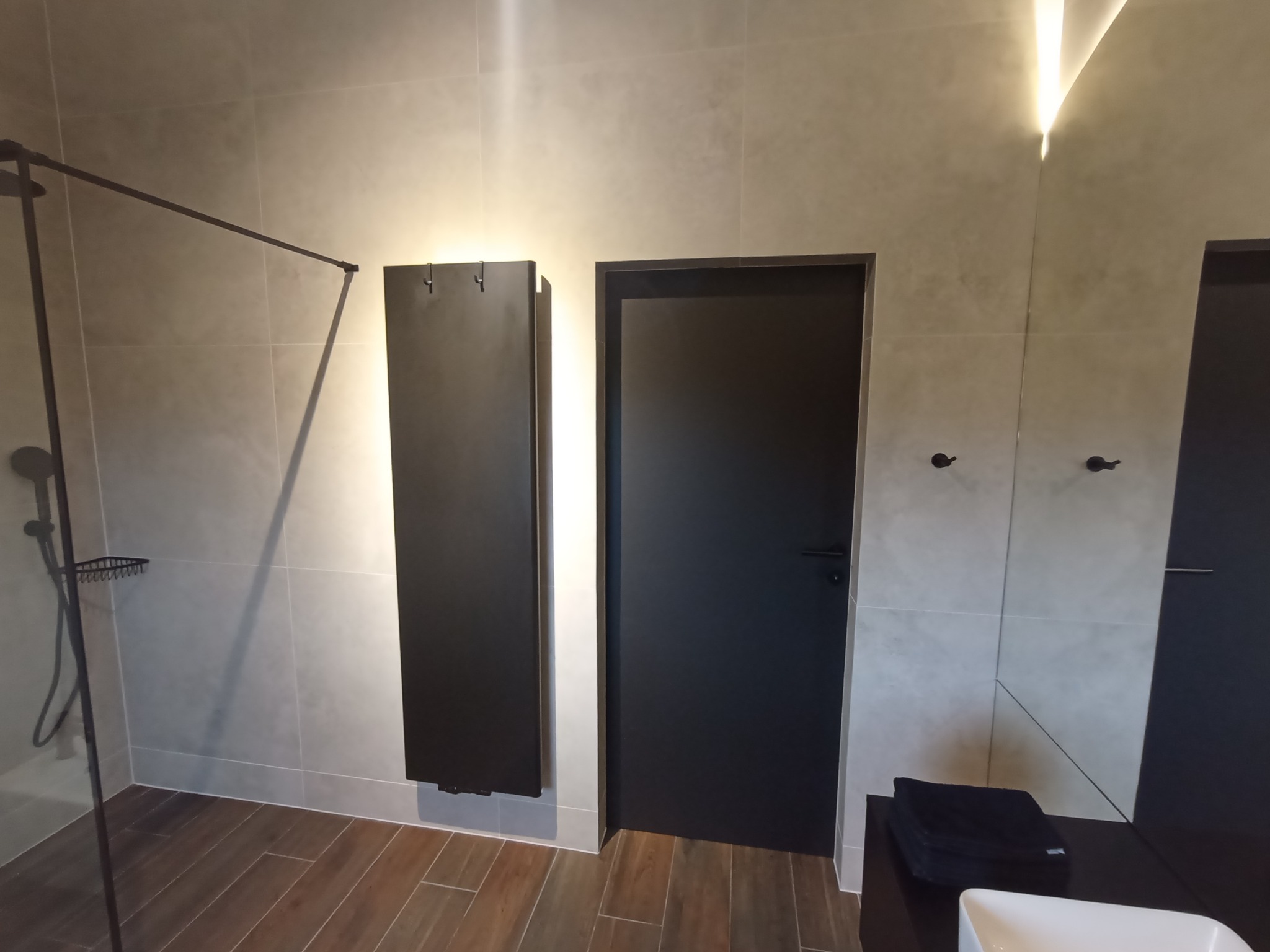 Nowoczesna łazienka z czarną armaturą, grzejnikiem drabinkowym i kabiną prysznicową typu walk-in, wykończona płytkami w odcieniach szarości i drewnopodobną podłogą.