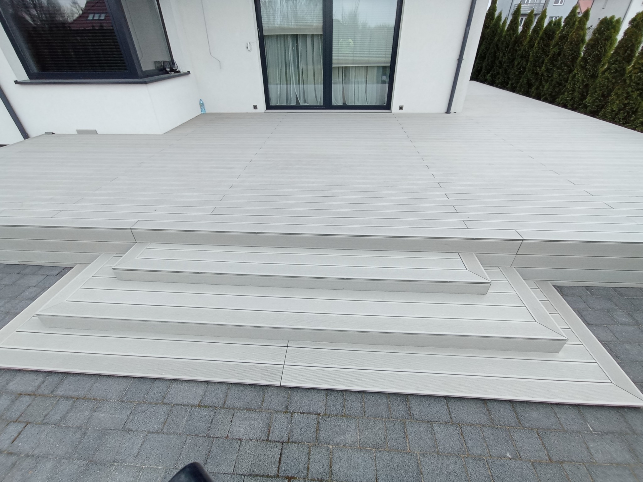 Nowoczesny taras z desek kompozytowych w jasnym odcieniu szarości, prowadzący do drzwi balkonowych, z widocznymi trzema stopniami i kostką brukową na pierwszym planie.