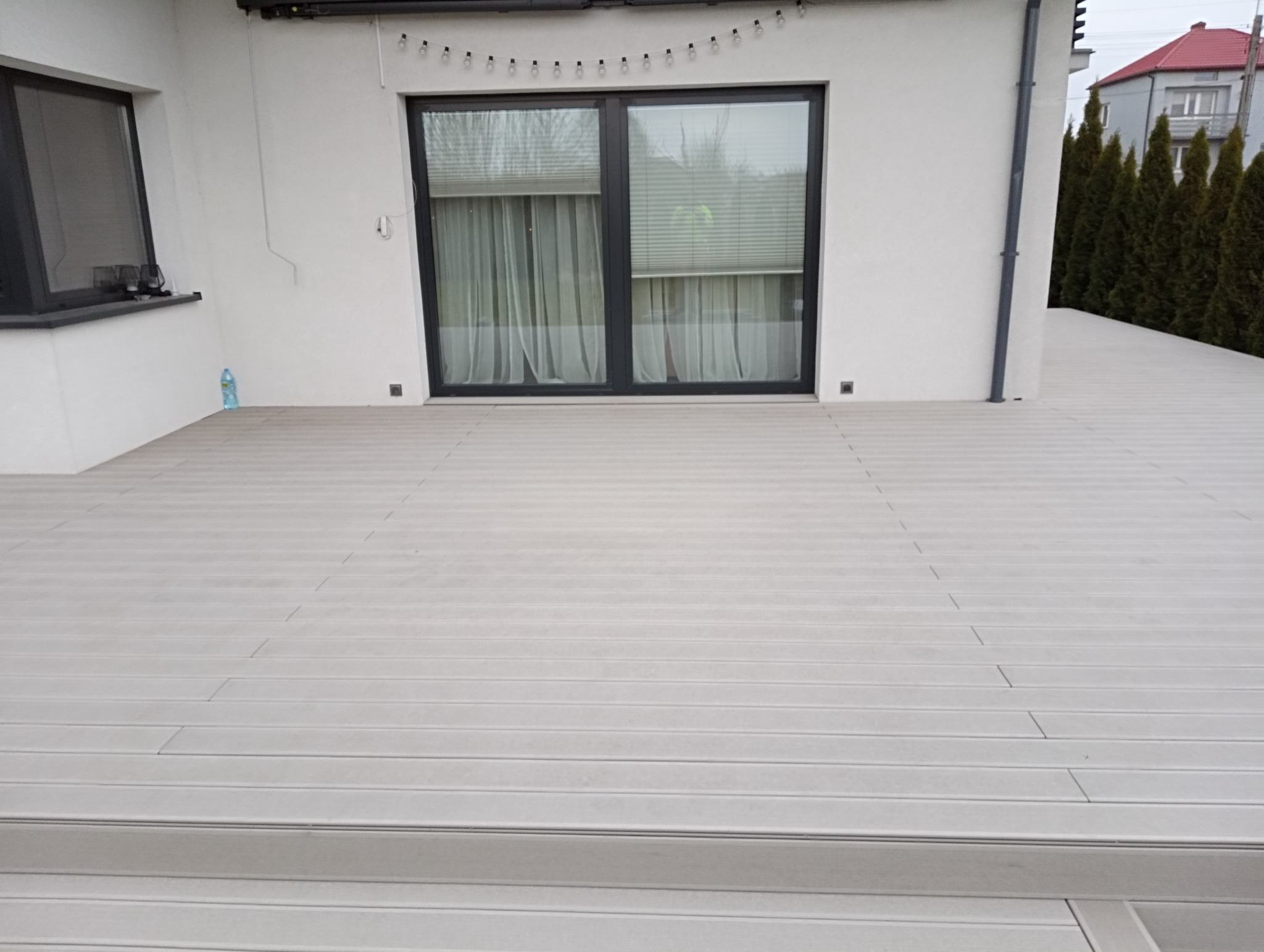 Jasnoszary taras kompozytowy z ryflowaną powierzchnią, przylegający do białego budynku z dużym oknem balkonowym i dekoracyjnymi lampkami na elewacji.