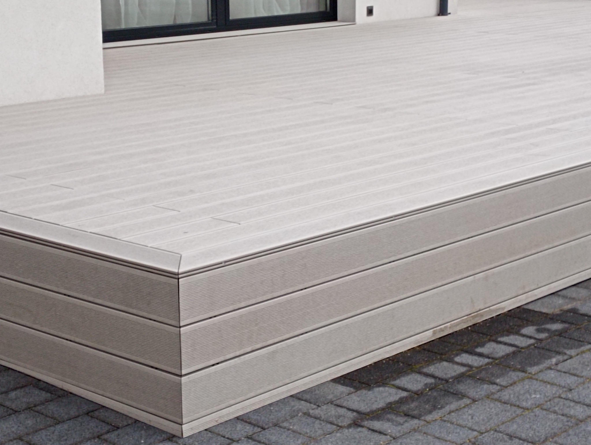 Nowoczesny taras z kompozytowych desek w odcieniach szarości, z widocznym detalem łączenia desek na narożniku i fragmentem elewacji budynku w tle.