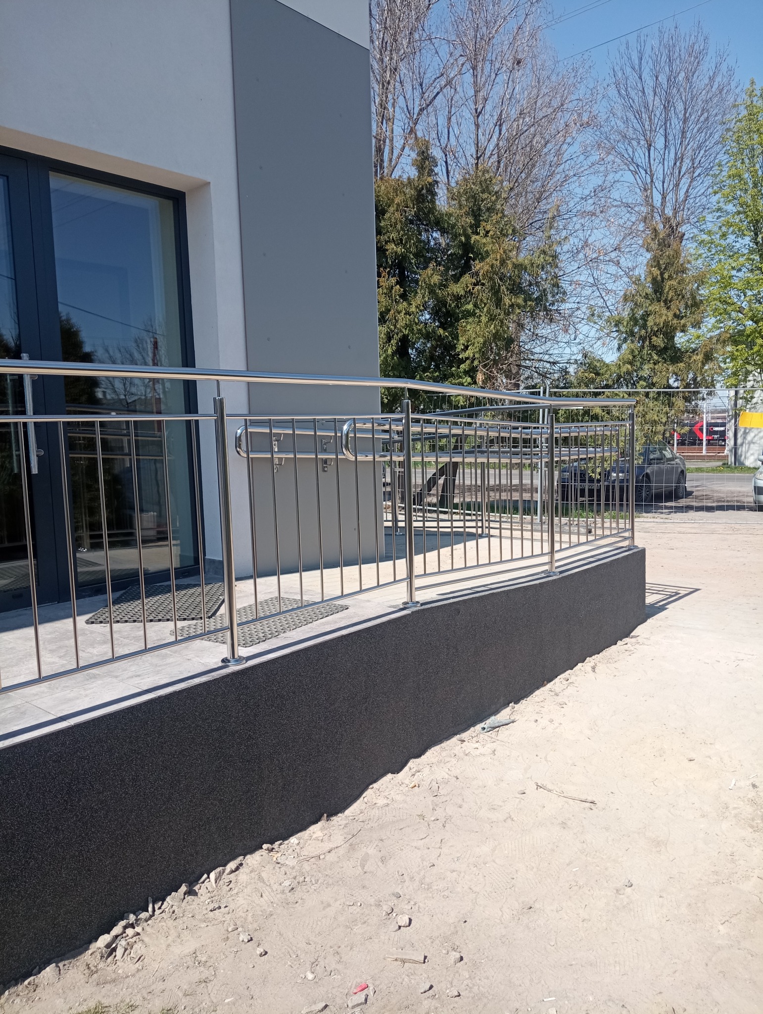 Zewnętrzna barierka ze stali nierdzewnej zamontowana przy wejściu do budynku z szarą elewacją i ciemną podmurówką, widok na drzewa i samochody w tle.