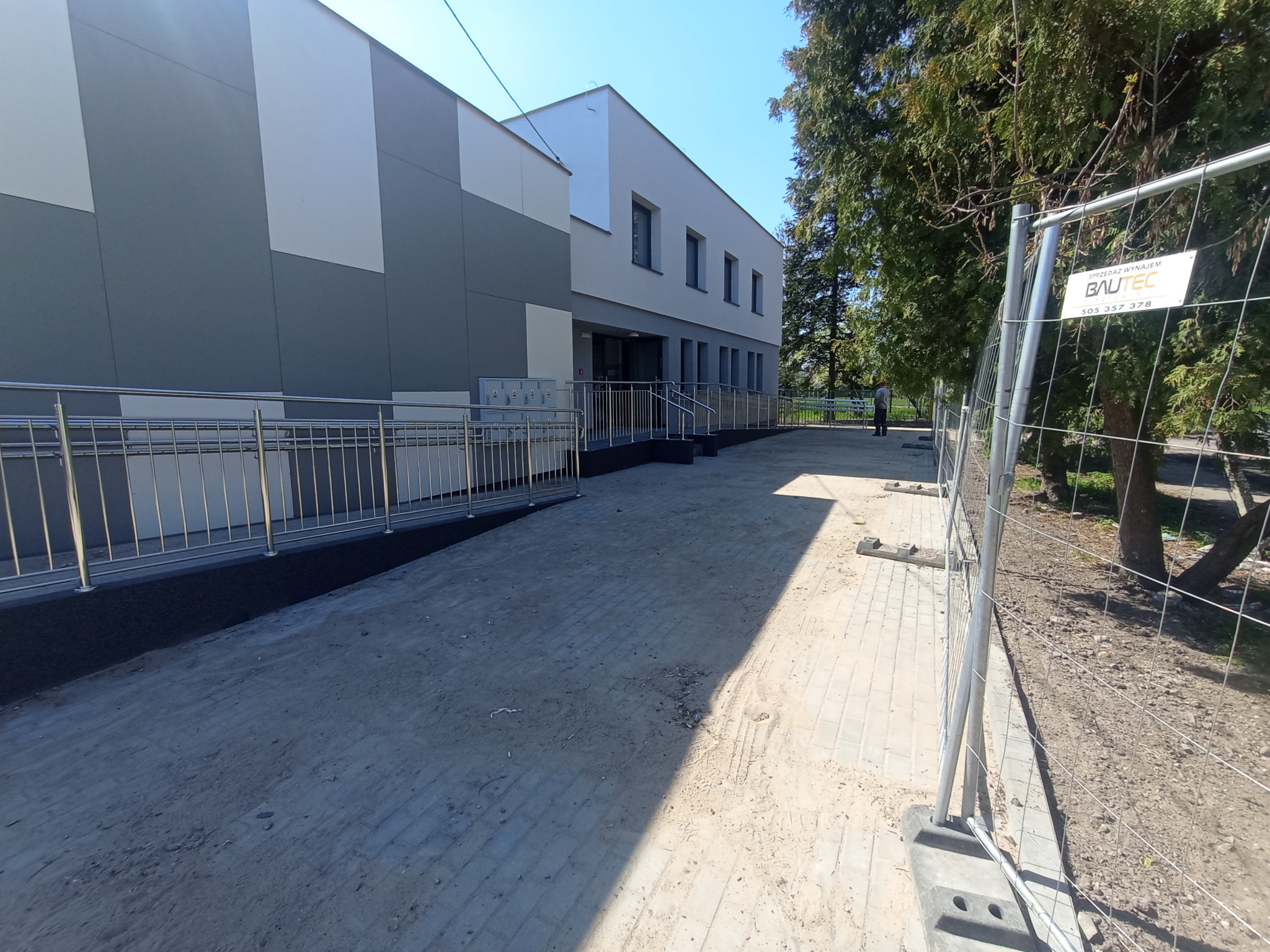 Nowoczesny budynek z szaro-białą elewacją, widoczny podjazd dla niepełnosprawnych z metalowymi barierkami, teren wokół w trakcie prac wykończeniowych, zabezpieczony tymczasowym ogrodzeniem...