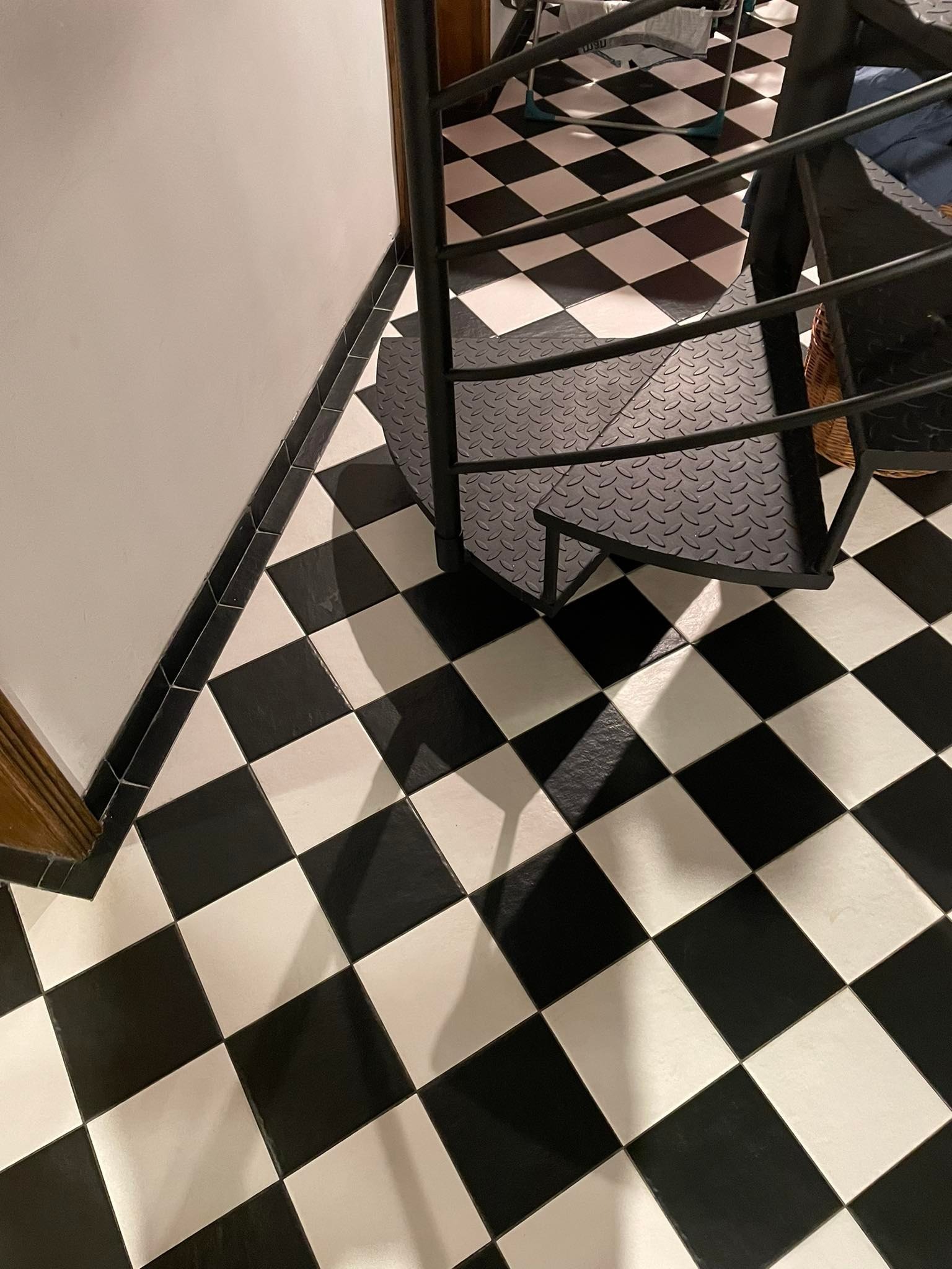 Czarno-biała podłoga w szachownicę z płytkami ceramicznymi, widok od góry, z fragmentem czarnej metalowej spiralnej klatki schodowej i czarnymi listwami przypodłogowymi.