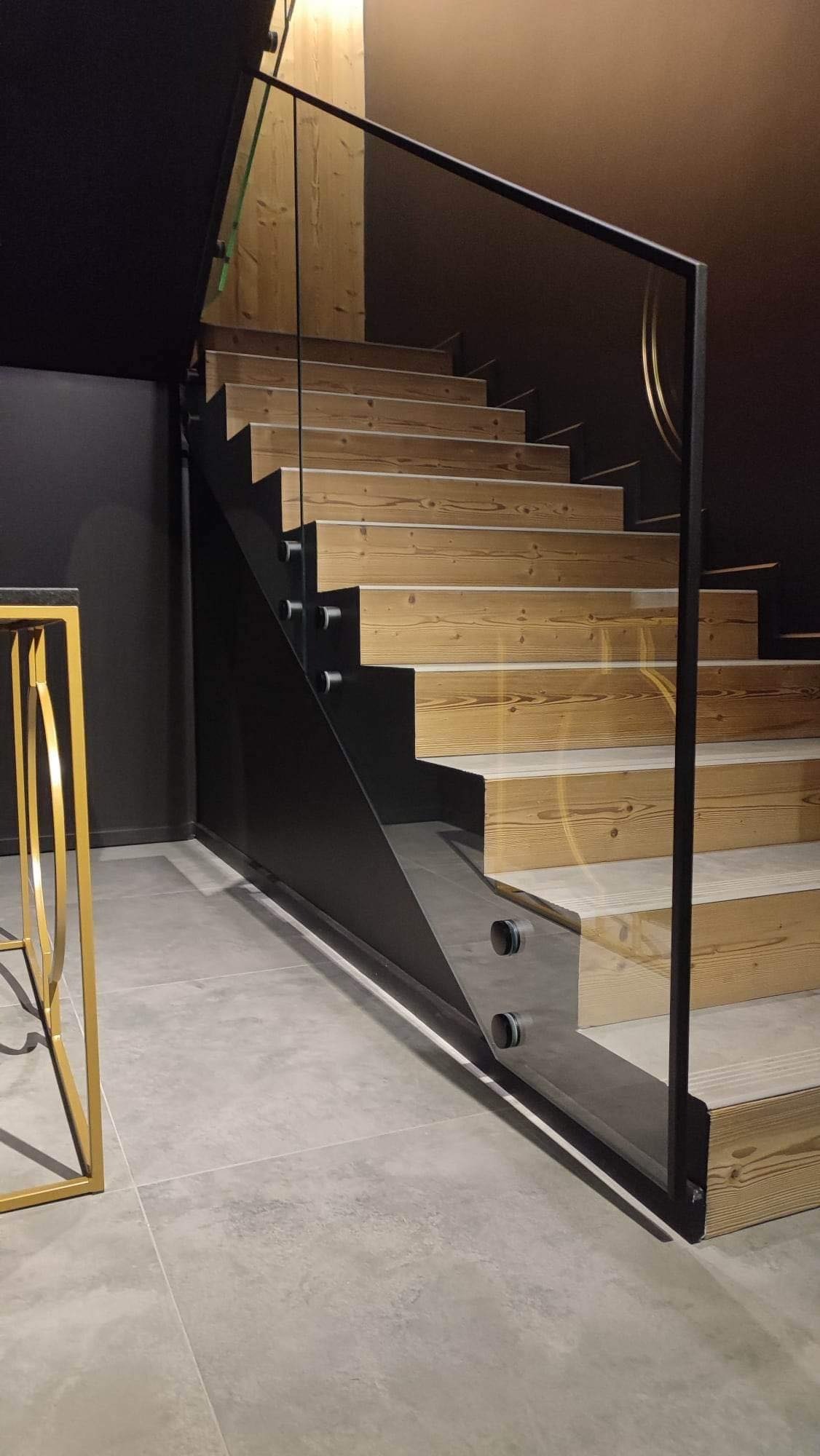 Nowoczesne schody drewniane z szklaną balustradą i czarnymi elementami metalowymi, oświetlenie górne, szare płytki podłogowe.