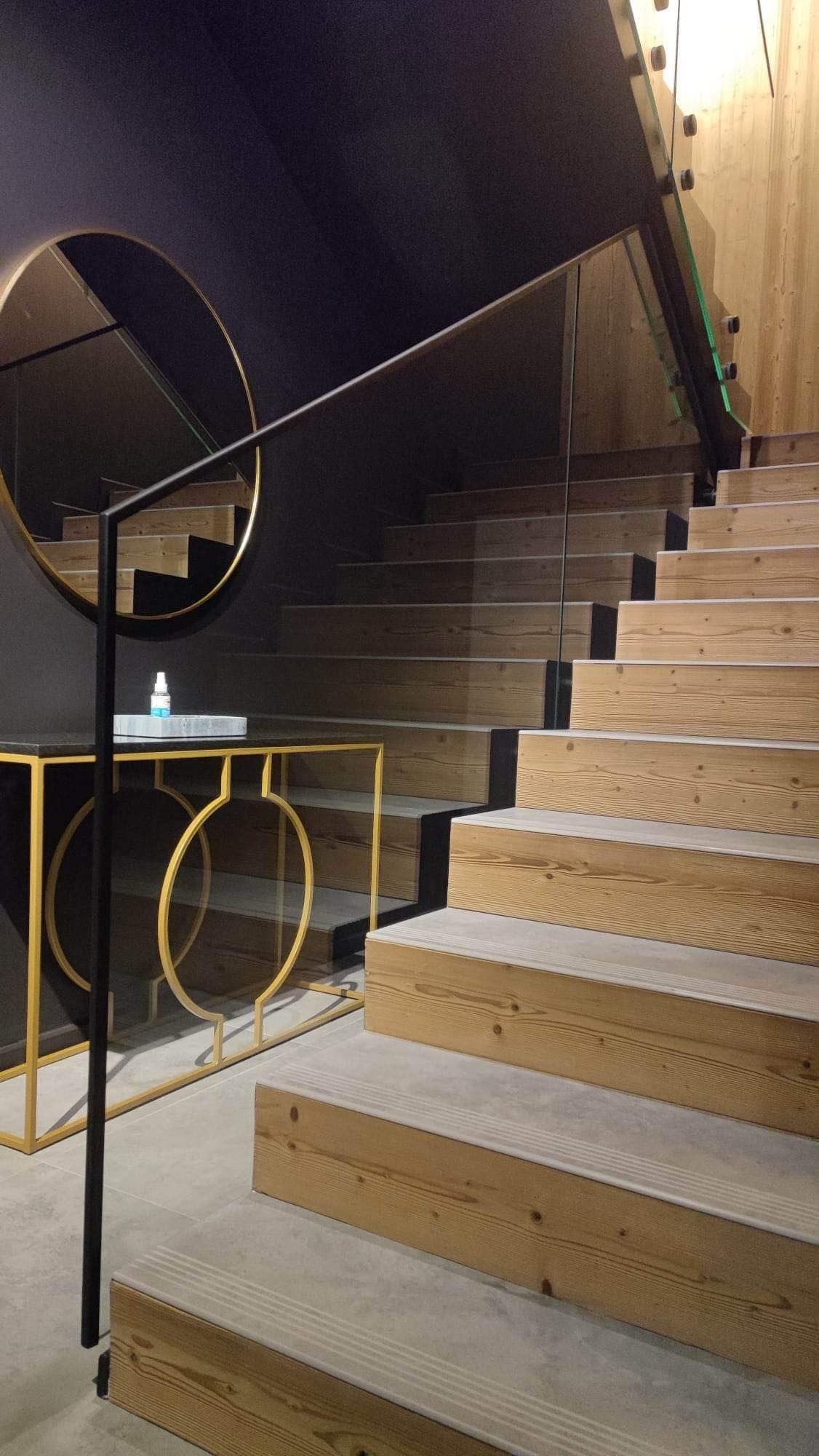 Nowoczesne drewniane schody z szarą wstawką, zabezpieczone szklaną balustradą, obok okrągłe lustro na czarnej ścianie i stolik z marmurowym blatem oraz złotą podstawą.