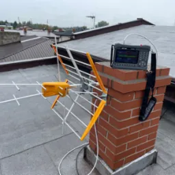 Antena telewizyjna przymocowana do komina na dachu, obok analizator sygnału SATLINK ST-6906 z podłączonym kablem, widok na okoliczne dachy w pochmurny dzień.