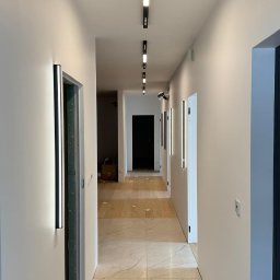 Qualitas Marek Zawadzki - Długi korytarz z nowoczesnym oświetleniem: podłużne lampy sufitowe z rzędem diod i pionowe, wąskie lampy ścienne, jasne ściany, marmurowe płytki podłogowe, widoczne drzwi i otwory drzwiowe.