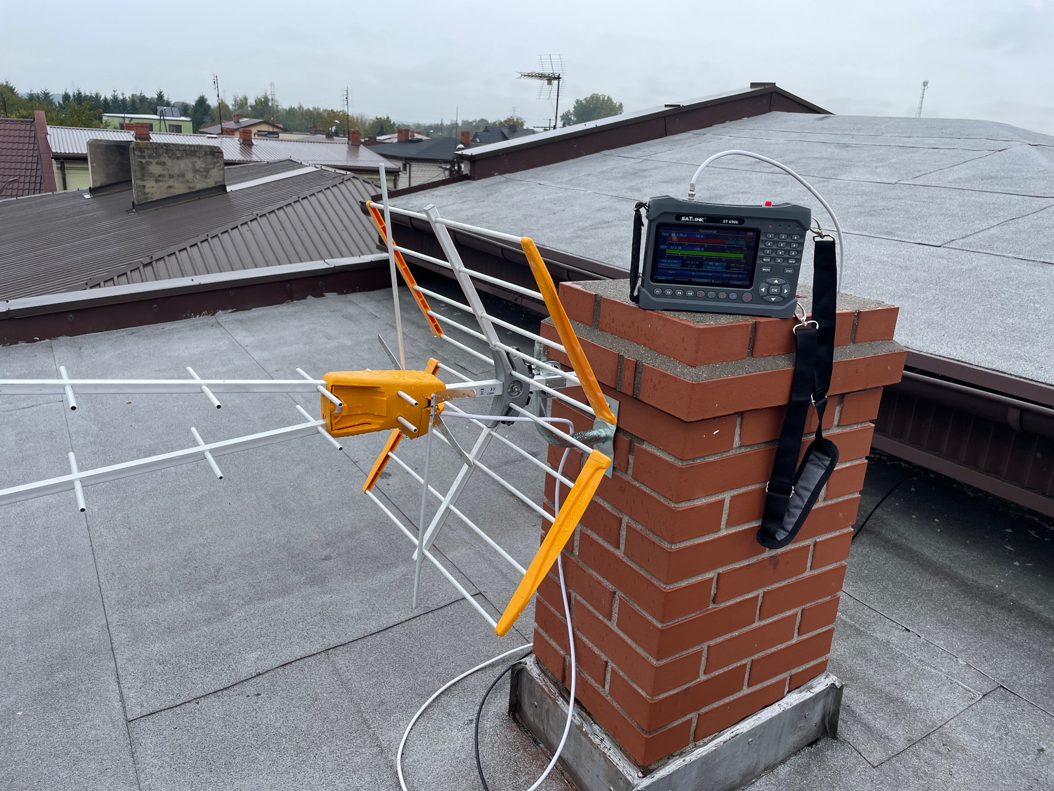 Antena telewizyjna przymocowana do komina na dachu, obok analizator sygnału SATLINK ST-6906 z podłączonym kablem, widok na okoliczne dachy w pochmurny dzień.