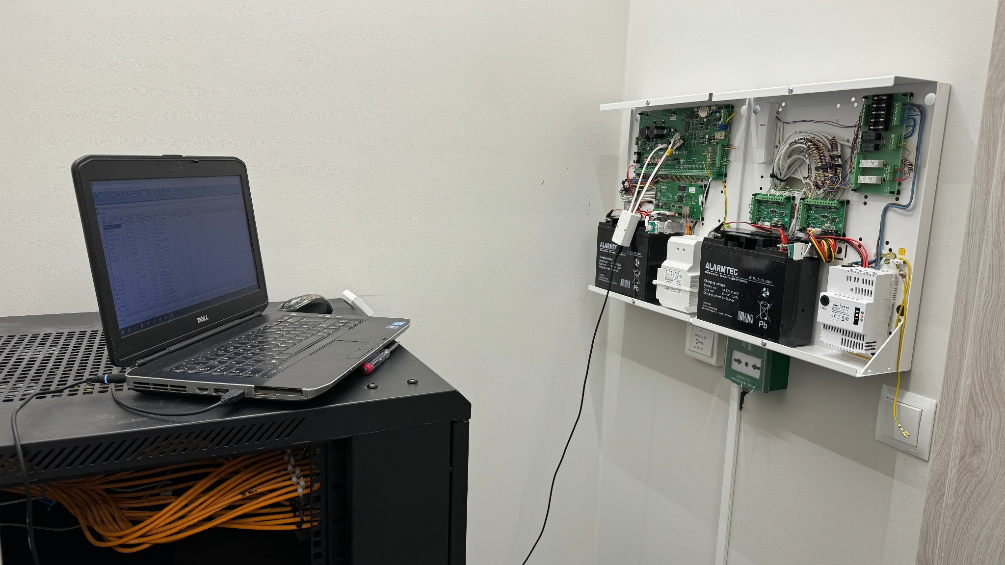 Konfiguracja systemu alarmowego w Koszalinie: Laptop podłączony do centrali alarmowej z akumulatorami i modułami, widoczne okablowanie w szafie rack.
