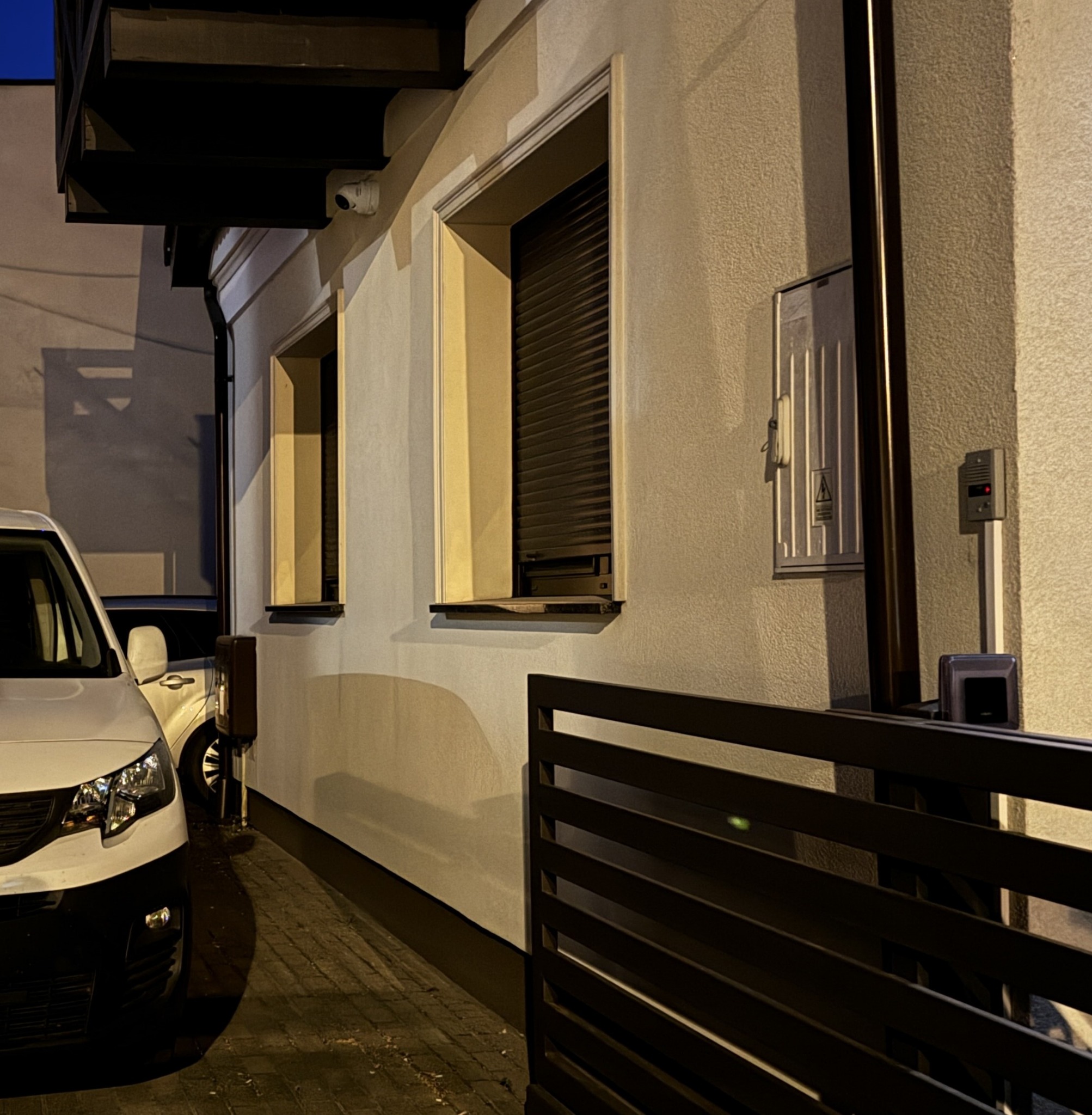 Zewnętrzny widok budynku z zainstalowaną kamerą monitoringu nad oknem z roletami, skrzynką elektryczną i domofonem, obok zaparkowany biały samochód dostawczy.