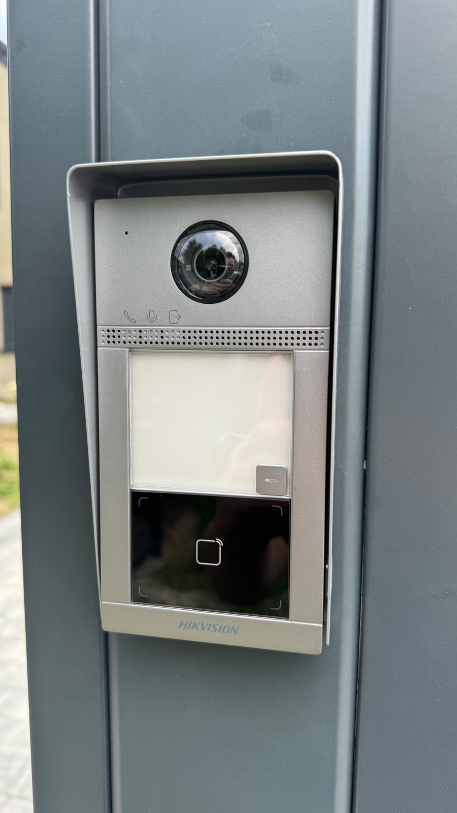 Szary wideodomofon Hikvision zamontowany na metalowej bramie, widoczna kamera, ekran i czytnik kart zbliżeniowych.