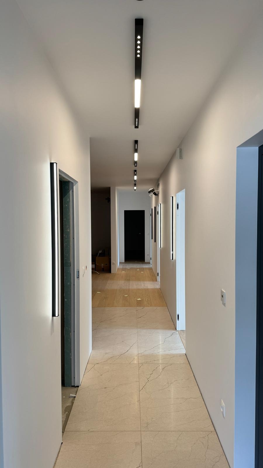 Długi korytarz z nowoczesnym oświetleniem: podłużne lampy sufitowe z rzędem diod i pionowe, wąskie lampy ścienne, jasne ściany, marmurowe płytki podłogowe, widoczne drzwi i otwory drzwiowe.