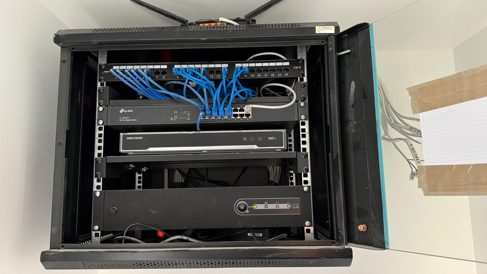 Szafa rack z zainstalowanymi urządzeniami sieciowymi, w tym switch TP-Link, rejestrator Hikvision i patch panel z niebieskimi kablami Ethernet. Widoczne okablowanie wychodzące z boku szafy.
