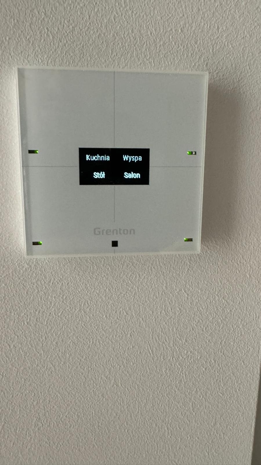 Biały panel sterowania systemem inteligentnego domu marki Grenton, zamontowany na teksturowanej ścianie, z podświetlanymi na zielono diodami i wyświetlaczem wskazującym nazwy pomieszczeń: Kuchnia...
